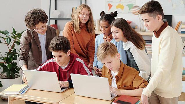 Mehrere Kinder sitzen und stehen hinter einem Tisch mit zwei Laptops.