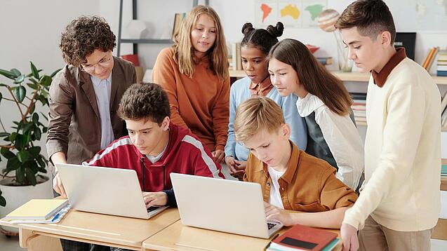 Mehrere Kinder sitzen und stehen hinter einem Tisch mit zwei Laptops.