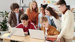 Mehrere Kinder sitzen und stehen hinter einem Tisch mit zwei Laptops.