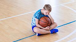 Ein Junge sitzt niedergeschlagen auf dem Boden einer Turnhalle und hält einen Basketball.