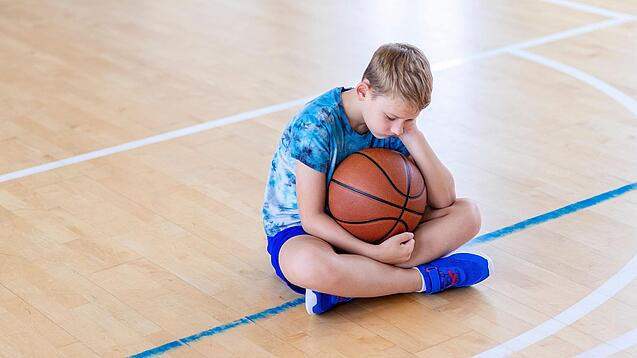 Ein Junge sitzt niedergeschlagen auf dem Boden einer Turnhalle und hält einen Basketball.