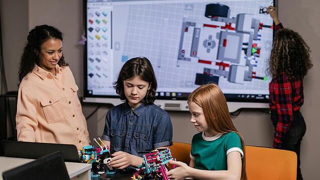 Zwei Schüler basteln an einem Lego-Roboter. Die Lehrerin steht daneben und schaut zu. Eine Schülerin schreibt im Hintergrund auf ein Whiteboard.