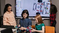 Zwei Schüler basteln an einem Lego-Roboter. Die Lehrerin steht daneben und schaut zu. Eine Schülerin schreibt im Hintergrund auf ein Whiteboard.