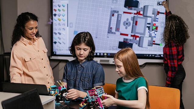 Zwei Schüler basteln an einem Lego-Roboter. Die Lehrerin steht daneben und schaut zu. Eine Schülerin schreibt im Hintergrund auf ein Whiteboard.