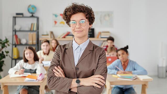 Eine Lehrerin mit Brille steht mit verschränkten Armen vor einer Schulklasse
