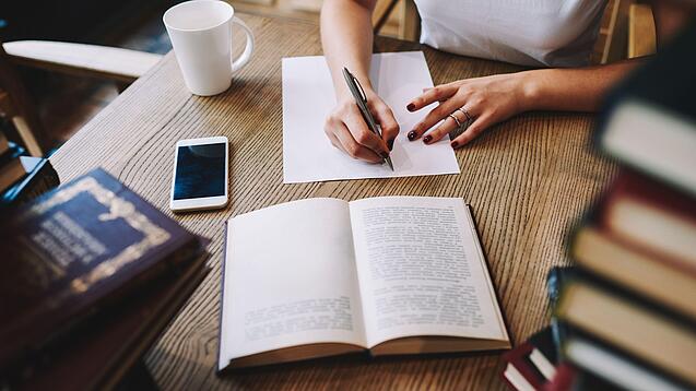 Eine Person sitzt an einem Tisch, Bücher, ein Handy und eine Tasse um sich herum und schreibt etwas auf.