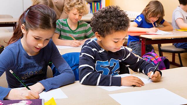 Grundschüler sitzen an Tischen in einem Klassenraum und schreiben auf einem Blatt Papier.