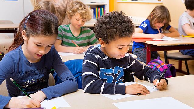 Grundschüler sitzen an Tischen in einem Klassenraum und schreiben auf einem Blatt Papier.