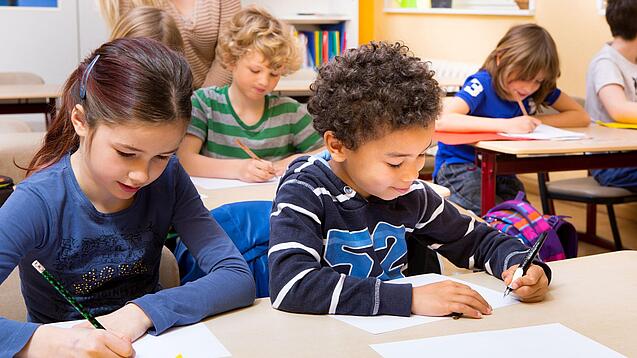 Grundschüler sitzen an Tischen in einem Klassenraum und schreiben auf einem Blatt Papier.