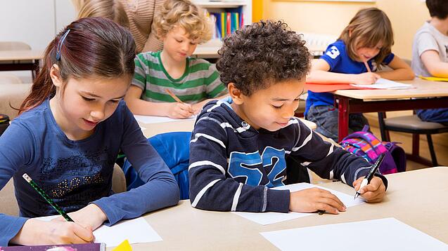 Grundschüler sitzen an Tischen in einem Klassenraum und schreiben auf einem Blatt Papier.