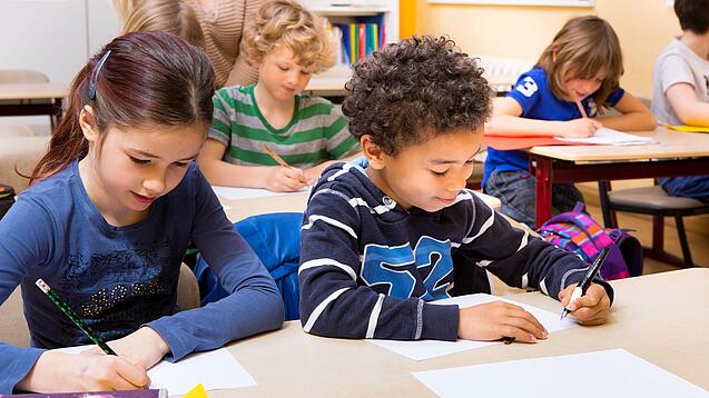 Grundschüler sitzen an Tischen in einem Klassenraum und schreiben auf einem Blatt Papier.