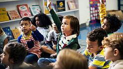 Kinder sitzen in einem Klassenraum auf dem Boden und zeigen auf.