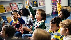 Kinder sitzen in einem Klassenraum auf dem Boden und zeigen auf.