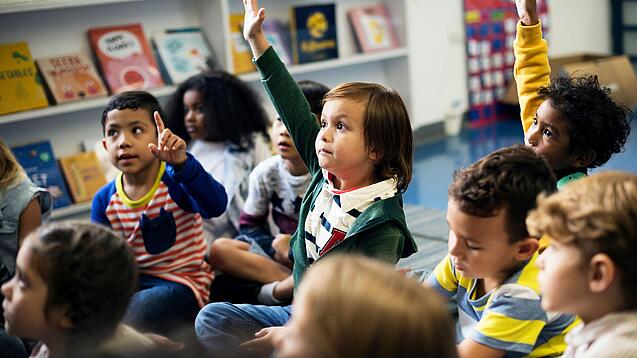 Kinder sitzen in einem Klassenraum auf dem Boden und zeigen auf.