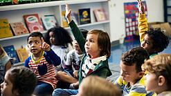Kinder sitzen in einem Klassenraum auf dem Boden und zeigen auf.