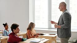 Ein Lehrer steht vor einer Klasse und erklärt etwas; die Schüler:innen hören konzentriert zu.