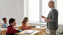 Ein Lehrer steht vor einer Klasse und erklärt etwas; die Schüler:innen hören konzentriert zu.