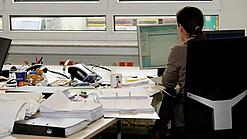 Eine Frau sitzt an einem Schreibtisch mit vielen Unterlagen und schaut auf einen Computer. 