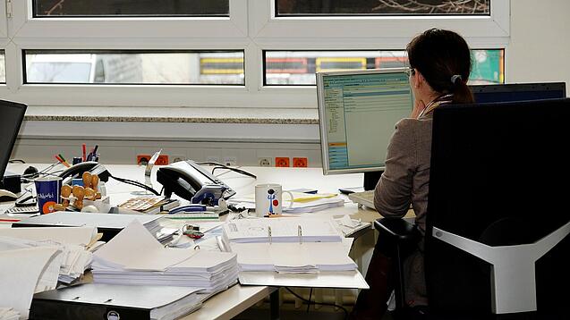 Eine Frau sitzt an einem Schreibtisch mit vielen Unterlagen und schaut auf einen Computer. 