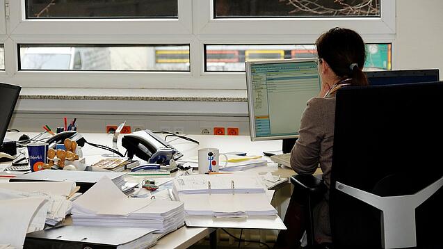 Eine Frau sitzt an einem Schreibtisch mit vielen Unterlagen und schaut auf einen Computer. 