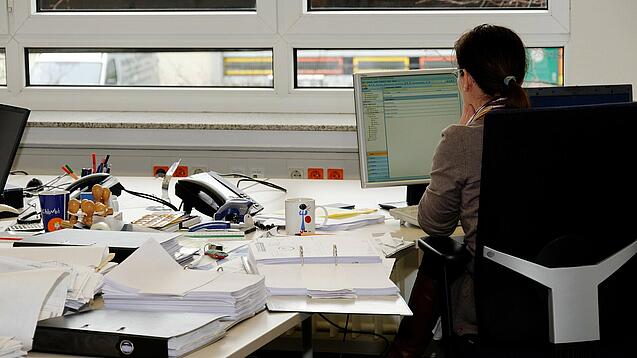 Eine Frau sitzt an einem Schreibtisch mit vielen Unterlagen und schaut auf einen Computer. 