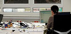 Eine Frau sitzt an einem Schreibtisch mit vielen Unterlagen und schaut auf einen Computer. 