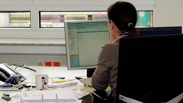 Eine Frau sitzt an einem Schreibtisch mit vielen Unterlagen und schaut auf einen Computer. 