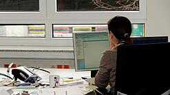 Eine Frau sitzt an einem Schreibtisch mit vielen Unterlagen und schaut auf einen Computer. 