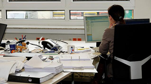 Eine Frau sitzt an einem Schreibtisch mit vielen Unterlagen und schaut auf einen Computer. 
