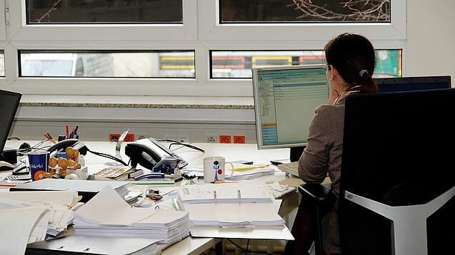 Eine Frau sitzt an einem Schreibtisch mit vielen Unterlagen und schaut auf einen Computer. 