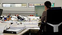 Eine Frau sitzt an einem Schreibtisch mit vielen Unterlagen und schaut auf einen Computer. 