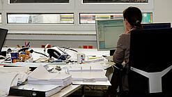 Eine Frau sitzt an einem Schreibtisch mit vielen Unterlagen und schaut auf einen Computer. 