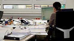 Eine Frau sitzt an einem Schreibtisch mit vielen Unterlagen und schaut auf einen Computer. 