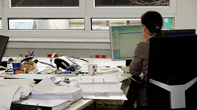 Eine Frau sitzt an einem Schreibtisch mit vielen Unterlagen und schaut auf einen Computer. 