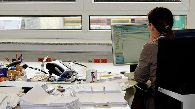 Eine Frau sitzt an einem Schreibtisch mit vielen Unterlagen und schaut auf einen Computer. 