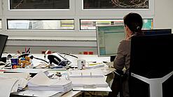 Eine Frau sitzt an einem Schreibtisch mit vielen Unterlagen und schaut auf einen Computer. 
