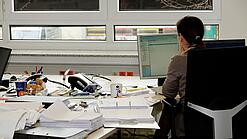 Eine Frau sitzt an einem Schreibtisch mit vielen Unterlagen und schaut auf einen Computer. 