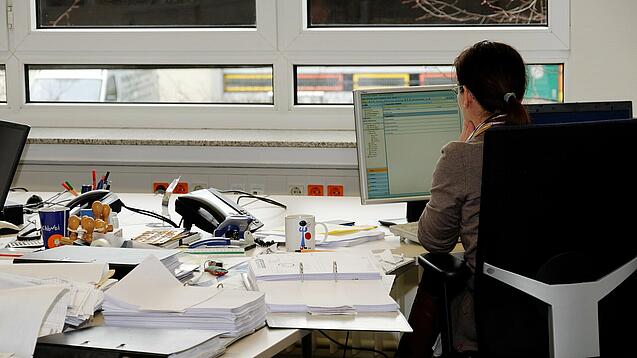 Eine Frau sitzt an einem Schreibtisch mit vielen Unterlagen und schaut auf einen Computer. 