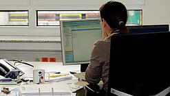 Eine Frau sitzt an einem Schreibtisch mit vielen Unterlagen und schaut auf einen Computer. 