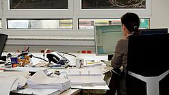 Eine Frau sitzt an einem Schreibtisch mit vielen Unterlagen und schaut auf einen Computer. 