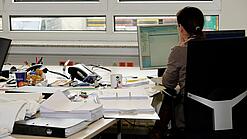 Eine Frau sitzt an einem Schreibtisch mit vielen Unterlagen und schaut auf einen Computer. 