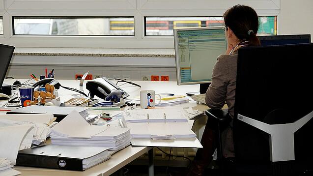Eine Frau sitzt an einem Schreibtisch mit vielen Unterlagen und schaut auf einen Computer. 