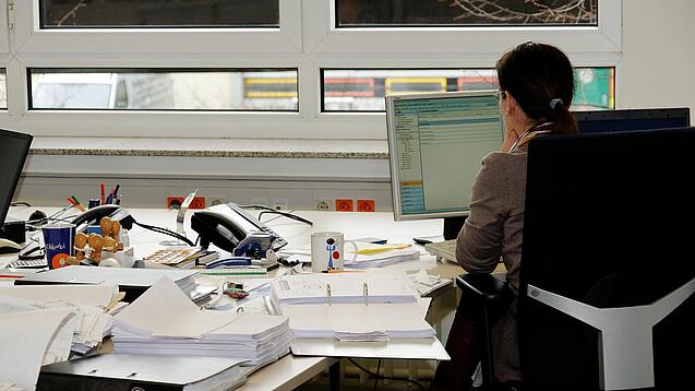 Eine Frau sitzt an einem Schreibtisch mit vielen Unterlagen und schaut auf einen Computer. 