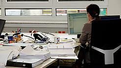 Eine Frau sitzt an einem Schreibtisch mit vielen Unterlagen und schaut auf einen Computer. 