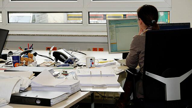 Eine Frau sitzt an einem Schreibtisch mit vielen Unterlagen und schaut auf einen Computer. 