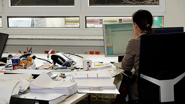 Eine Frau sitzt an einem Schreibtisch mit vielen Unterlagen und schaut auf einen Computer. 
