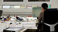 Eine Frau sitzt an einem Schreibtisch mit vielen Unterlagen und schaut auf einen Computer. 