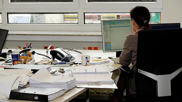 Eine Frau sitzt an einem Schreibtisch mit vielen Unterlagen und schaut auf einen Computer. 