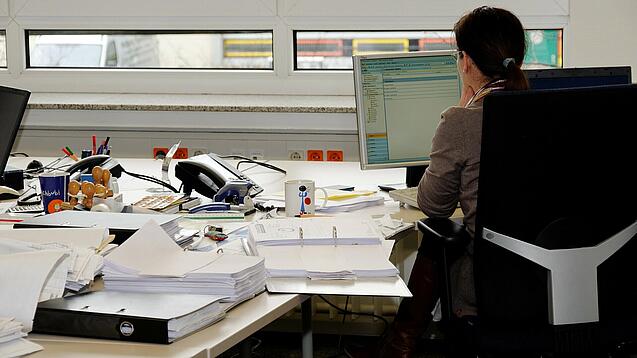 Eine Frau sitzt an einem Schreibtisch mit vielen Unterlagen und schaut auf einen Computer. 