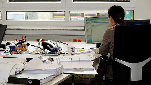 Eine Frau sitzt an einem Schreibtisch mit vielen Unterlagen und schaut auf einen Computer. 